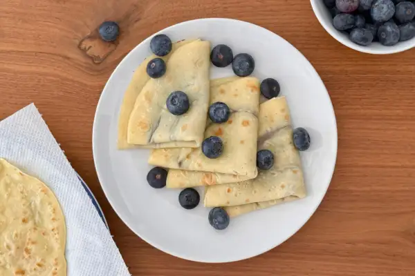 Deliziose crepes all'acqua, piegate e guarnite con mirtilli freschi, su un piatto bianco.