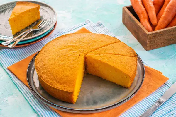 Una soffice torta di carote, appena affettata, su un piatto. Accanto, un piattino con una fetta e delle carote fresche in un cesto.