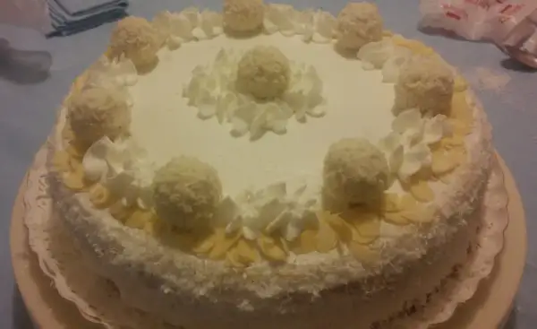 Torta Raffaello ricoperta di cocco, decorata con panna montata e praline, un'esplosione di dolcezza.
