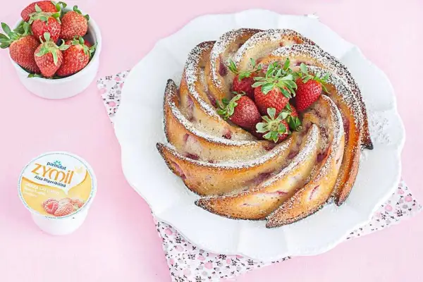 Torta di fragole e yogurt, soffice e profumata, decorata con zucchero a velo e fragole fresche. Accanto, una ciotolina di fragole e un vasetto di yogurt.