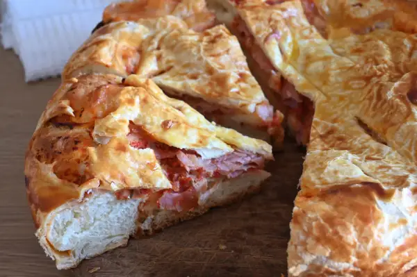 Una torta rustica con pasta sfoglia ripiena di salumi e pomodoro, tagliata a fette su un tagliere di legno.