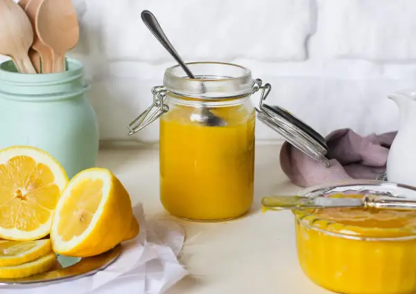 Vasetto di vetro pieno di lemon curd, con limoni freschi affettati e un cucchiaino.