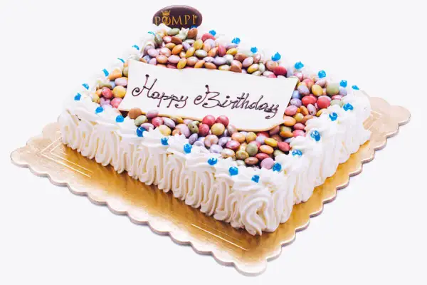 Torta rettangolare con glassa bianca, decorata con tanti smarties colorati e la scritta "Happy Birthday".