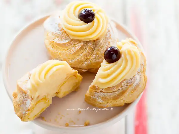 Zeppole di San Giuseppe, dolci soffici con crema pasticcera e ciliegina, uno è tagliato per mostrarne l'interno goloso.